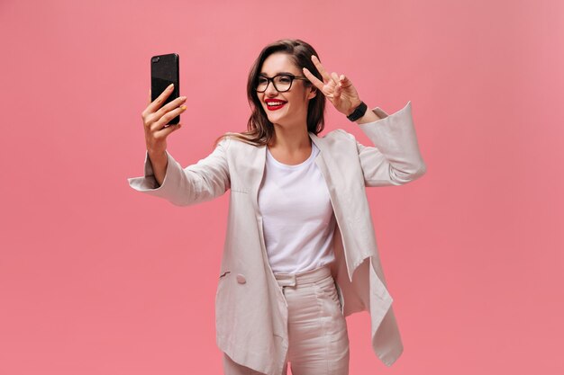 Merveilleuse jeune femme à lunettes élégantes et veste beige prend selfie et montre le signe de la paix sur fond isolé rose.