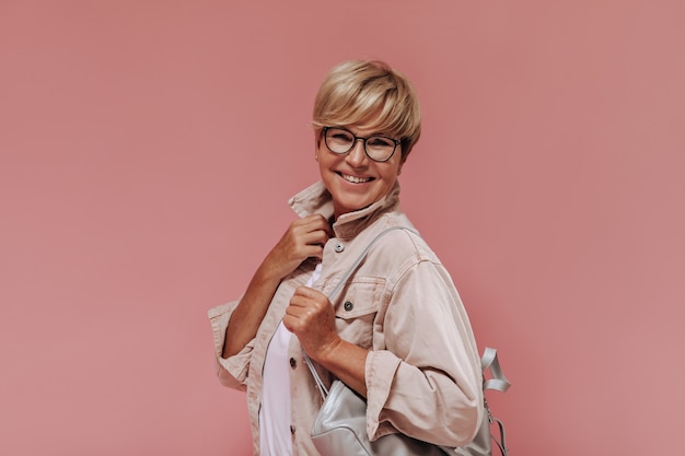 Merveilleuse vieille femme aux cheveux blonds et lunettes élégantes en veste beige et t-shirt léger souriant et posant avec sac sur fond rose.