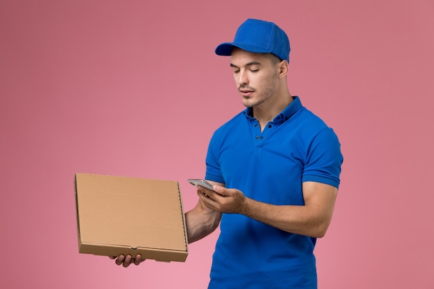 Messager mâle en uniforme bleu tenant la boîte de nourriture de livraison à l'aide de son téléphone sur rose, prestation de services de travail uniforme