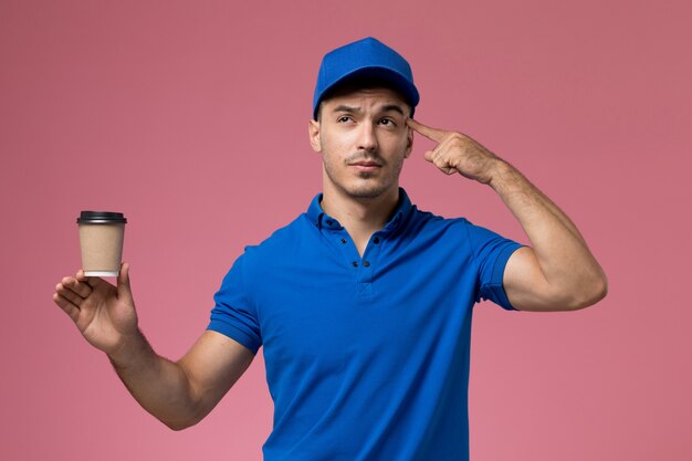 Messager masculin en uniforme bleu tenant la tasse de café de livraison en pensant à la prestation de services de travail rose, uniforme