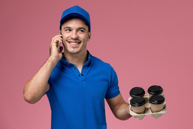 Messager masculin en uniforme bleu tenant des tasses à café et parler au téléphone sur rose, prestation de service uniforme