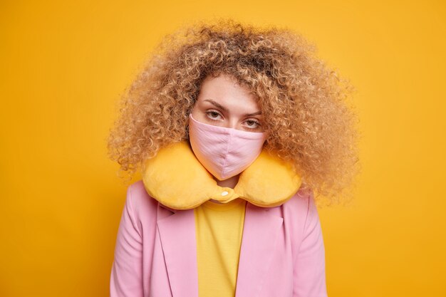 Mesures de sécurité pendant l'épidémie de virus. Une femme sérieuse a l'air avec confiance et porte un masque de protection pour faire correspondre les vêtements posés par l'oreiller au cou contre un mur jaune vif. Confinement et covid 19