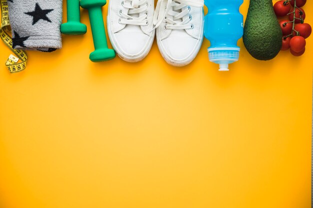 Mètre ruban; brassard; des haltères; chaussures; bouteille d'eau avocat et tomates cerises sur fond jaune