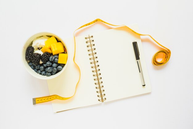 Mètre ruban; verre de fruits avec du yaourt; stylo et cahier à spirale sur fond blanc