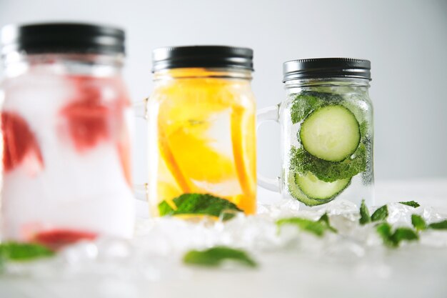 Mettre l'accent sur le dernier pot Limonades maison fraîches et saines avec de l'eau pétillante fraise, concombre, menthe et orange isolés dans des glaçons écrasés sur une table en bois