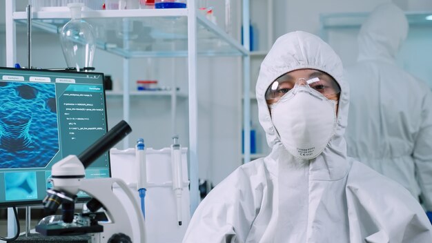 Microbiologiste assis dans un laboratoire portant un costume ppe regardant la caméra dans un laboratoire moderne équipé. Équipe de scientifiques examinant l'évolution du virus à l'aide d'outils de haute technologie et de chimie pour la recherche scientifique