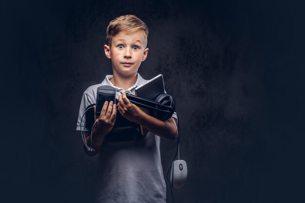 Mignon écolier vêtu d'un t-shirt blanc, contient un ensemble numérique complet pour le divertissement dans un studio. Isolé sur un fond texturé sombre.