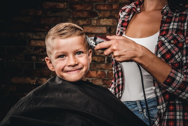 Photo gratuite mignon garçon d'âge préscolaire se coupe les cheveux. le coiffeur pour enfants avec tondeuse coupe un petit garçon dans la chambre avec un intérieur loft.