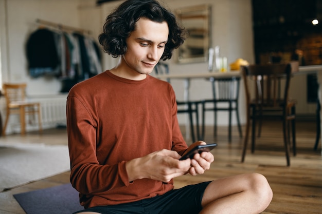 Mignon jeune étudiant aux cheveux ondulés assis sur le sol à la maison avec un téléphone portable en tapant un message texte à des amis via un chat de groupe sur Messenger, en regardant à travers les mèmes, en parcourant son compte de réseau social