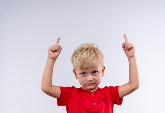 Un mignon petit garçon aux cheveux blonds et aux yeux bleus portant un t-shirt rouge pointant vers le haut avec l'index à côté sur un mur blanc