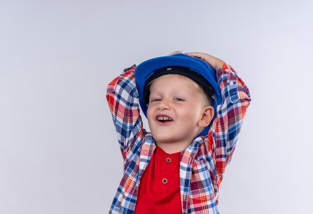 Un mignon petit garçon aux cheveux blonds portant chemise à carreaux en casque bleu tenant les mains sur sa tête sur un mur blanc