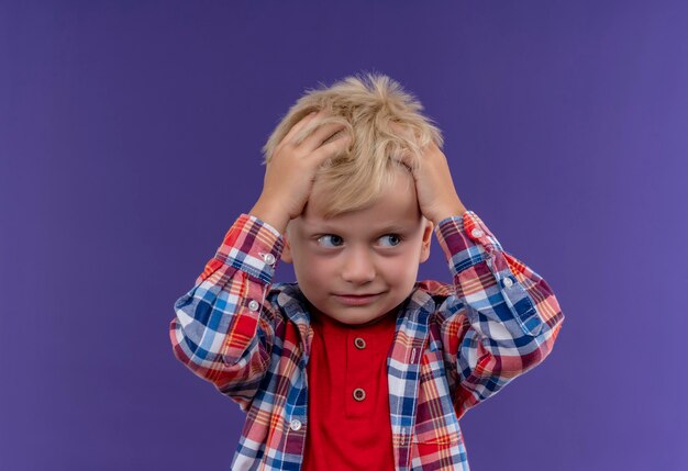 Un mignon petit garçon aux cheveux blonds portant une chemise à carreaux en gardant la main sur sa tête sur un mur violet