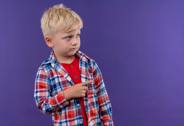 Un mignon petit garçon aux cheveux blonds portant chemise à carreaux pointant avec l'index sur quelque chose sur un mur violet