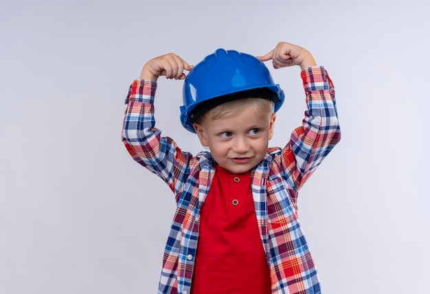 Un mignon petit garçon aux cheveux blonds portant chemise à carreaux pointant avec l'index sur son casque bleu sur un mur blanc