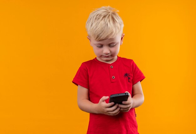 Un mignon petit garçon aux cheveux blonds portant un t-shirt rouge à la recherche de téléphone mobile sur un mur jaune