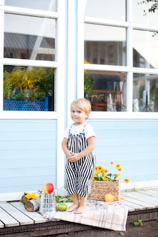 Un mignon petit garçon blond en salopette et t-shirt blanc se tient sur le porche d'une maison de campagne en bois un jour d'automne. reposez-vous dans la nature. concept d'enfance. récolte. petit fermier. mode de vie familial sain