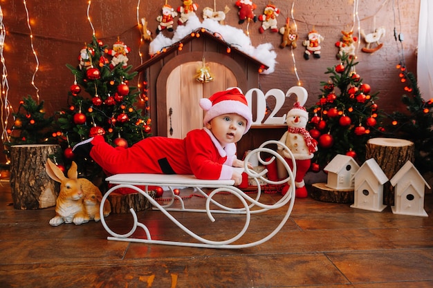 Mignon petit garçon habillé en père noël sur un vieux traîneau vintage avec des cadeaux près de l'arbre de noël, concept de joyeuses fêtes. 2022
