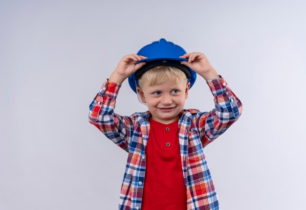 Un mignon petit garçon souriant aux cheveux blonds portant chemise à carreaux tenant son casque bleu avec les mains sur la tête sur un mur blanc