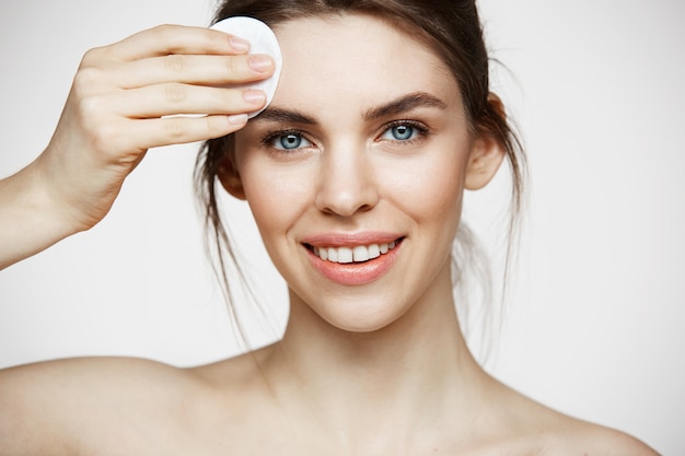 Mignonne belle fille brune naturelle, nettoyer le visage avec une éponge de coton souriant en regardant la caméra sur fond blanc. Cosmétologie et spa.