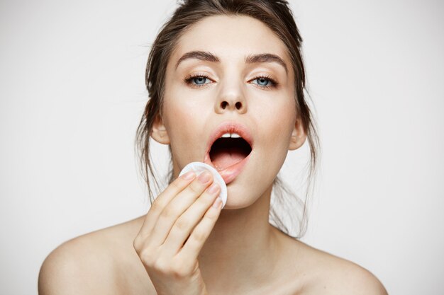 Mignonne belle fille brune naturelle, nettoyer le visage avec une éponge de coton souriant en regardant la caméra sur fond blanc. Cosmétologie et spa.