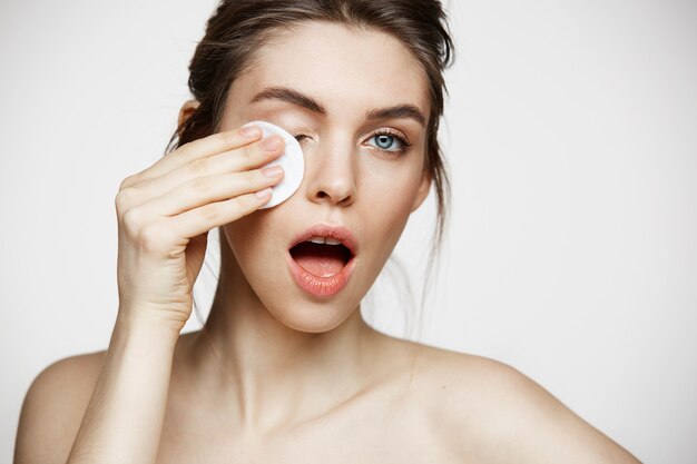 Mignonne belle fille brune naturelle, nettoyer le visage avec une éponge de coton souriant en regardant la caméra sur fond blanc. Cosmétologie et spa.