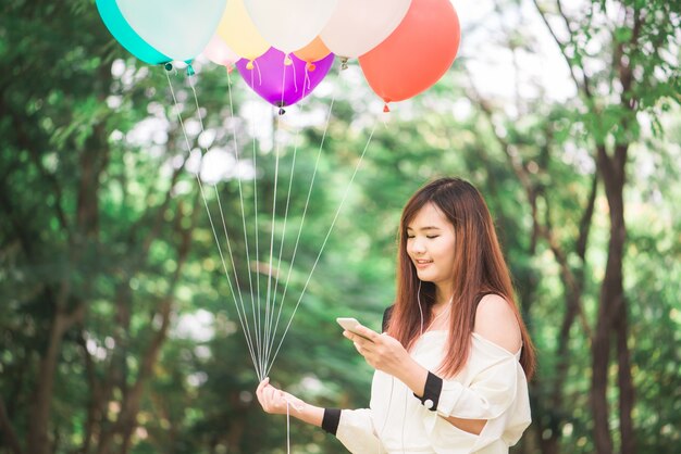 Mignonne femme asiatique est la lecture de message texte agréable sur un téléphone mobile tout en étant assis dans le parc au jour de printemps chaud, magnifique femme écoutant de la musique dans les écouteurs et la recherche d&#39;informations sur le téléphone cellulaire.