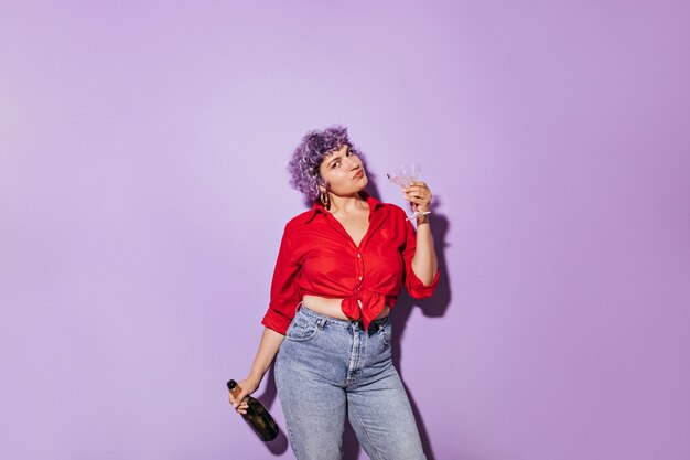 Mignonne femme merveilleuse en chemise élégante rouge détient verre et bouteille de vin blanc et sur lilas.