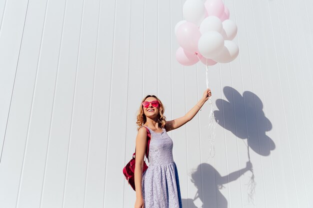 Mignonne jeune femme en lunettes de soleil roses appréciant la journée ensoleillée, tenant des ballons à air chaud