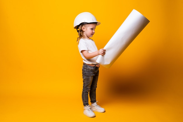 Mignonne petite fille ingénieur avec casque tenant bluepring isolé sur mur jaune