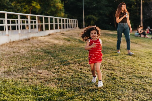 Mignonne petite fille en tenue rouge en cours d'exécution dans le parc. Charmant enfant aux cheveux bouclés jouant sur la pelouse.