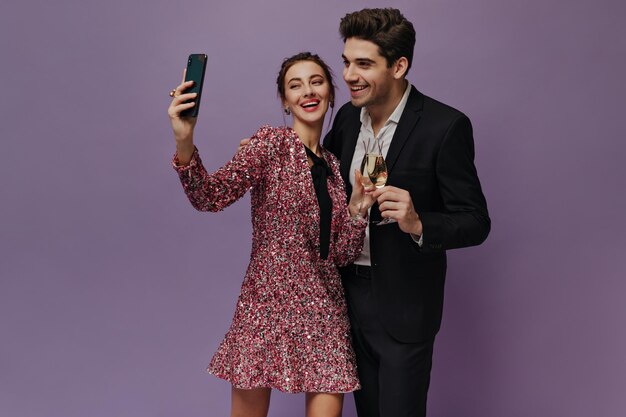 Mignons jeunes en tenue de fête souriant tenant des verres avec champagne et chat vidéo Paire posant isolé sur fond violet