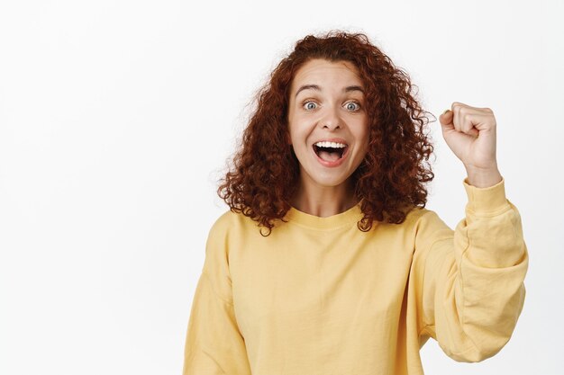 Une militante enthousiaste acclamant, stimulant la motivation en chantant, faisant un geste de pompe de poing et souriant émerveillée, debout en sweat-shirt jaune sur blanc.