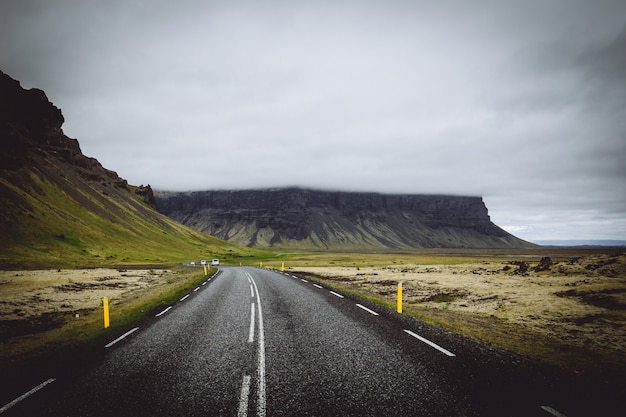 Une mince route dans un champ vert avec des collines et un ciel nuageux gris en Islande
