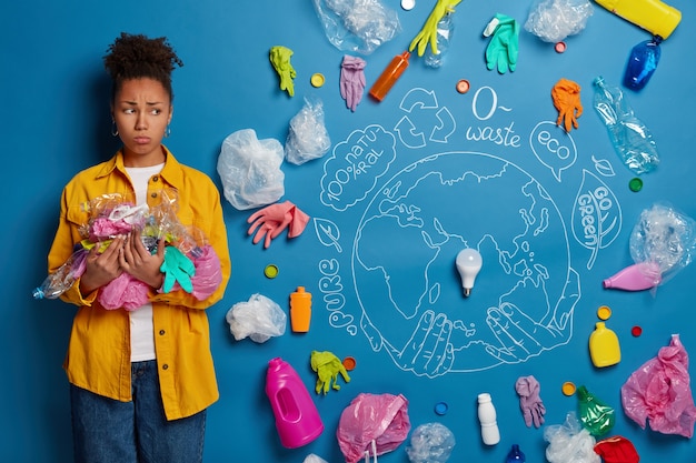 Misérable activiste bénévole de l'environnement à la peau sombre pose avec des déchets en plastique, ramasse les ordures, bouleversé comme vit sur une planète polluée, pose sur fond bleu.