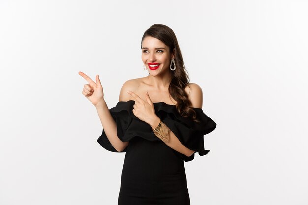 Mode et beauté. Jolie femme en bijoux, maquillage et robe noire, riant et pointant du doigt à gauche à l'offre promo, fond blanc.