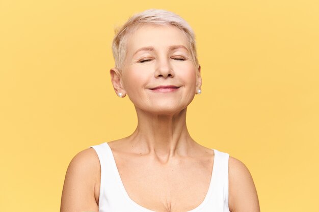 À la mode, belle femme de race blanche à la retraite avec une coiffure de lutin portant des vêtements décontractés posant en gardant les yeux fermés et souriant avec plaisir et plaisir, en écoutant de la bonne musique ou en rêvant