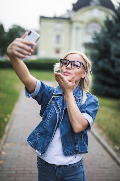 Mode élégante Fille Blonde Femme En Suite De Jeans Fait Selfie Donner Airkiss Sur Son Téléphone Dans La Ville Le Matin