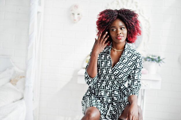 À la mode grande femme modèle afro-américaine avec des cheveux afro rouges en robe posée à la salle blanche assis sur une chaise contre le miroir