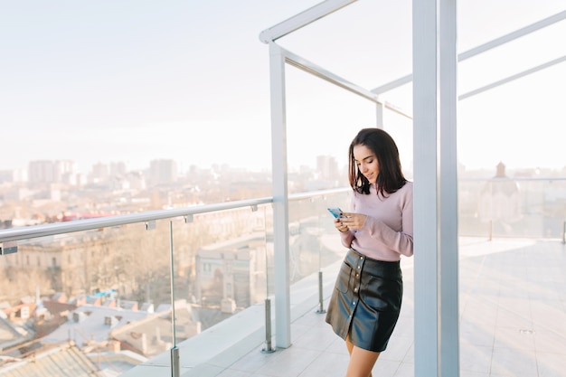 Mode jeune femme brune en jupe noire à l'aide de téléphone sur terrasse sur vue sur la ville.
