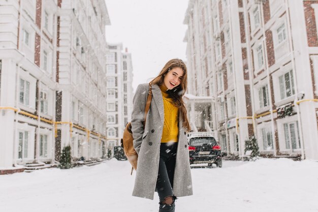Mode jeune femme en manteau avec sac à dos marchant sur la rue dans la grande ville en temps de neige. Humeur joyeuse, chutes de neige, attendant Noël, exprimant la positivité, les vraies émotions.