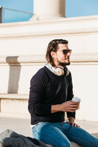 À la mode jeune homme avec un casque autour du cou, tenant une tasse de café à emporter