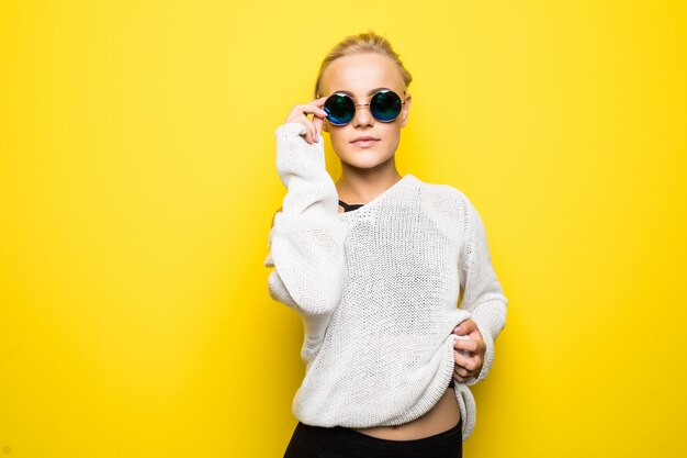 Mode jolie fille blonde en pull blanc moderne à lunettes de soleil bleu brillant pose sur jaune