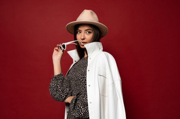 Mode portrait de belle femme brune maigre élégante avec un manteau blanc habillé et un chapeau beige tenant des lunettes de soleil. Style de mode tendance automne ou hiver.