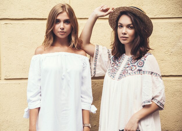 Mode portrait de deux jeunes femmes hippies élégantes brune et blonde modèles en journée ensoleillée d'été dans des vêtements blancs hipster posant près du mur jaune. Sans maquillage