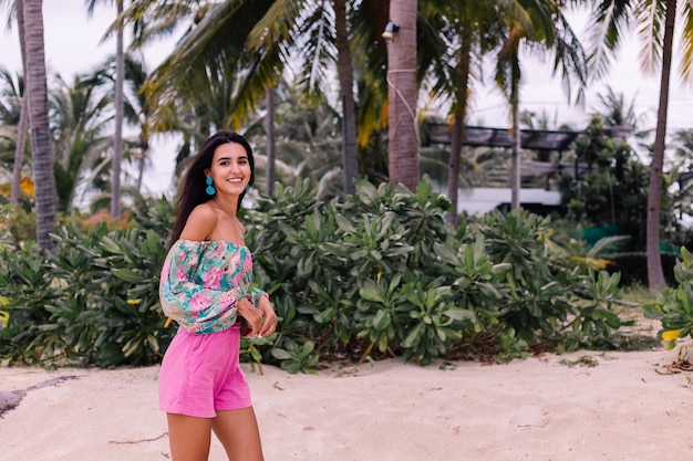 Mode portrait de femme élégante en haut à manches longues imprimé coloré et short rose sur la plage, fond tropical.