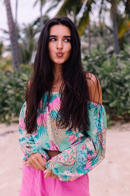 Mode portrait de femme élégante en haut à manches longues imprimé coloré et short rose sur la plage, fond tropical.