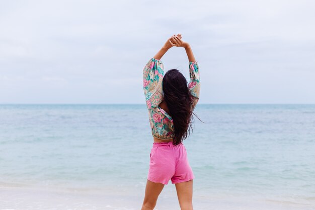 Mode portrait de femme élégante en haut à manches longues imprimé coloré et short rose sur la plage, fond tropical.