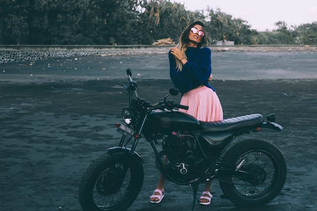 Photo gratuite mode portrait de femme élégante à lunettes de soleil, pull bleu et jupe rose, se dresse à vélo noir sur la plage de sable noir.