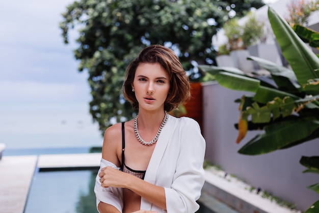 Mode portrait de femme à la villa de luxe tropicale portant un blazer élégant blanc et des bijoux sur les feuilles tropicales
