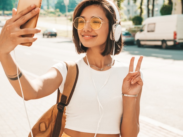 Mode portrait de jeune femme élégante hipster marchant dans la rue. Fille faisant selfie et montre le signe de la paix. Modèle souriant profitez de ses week-ends avec sac à dos. Femme écoutant de la musique via des écouteurs